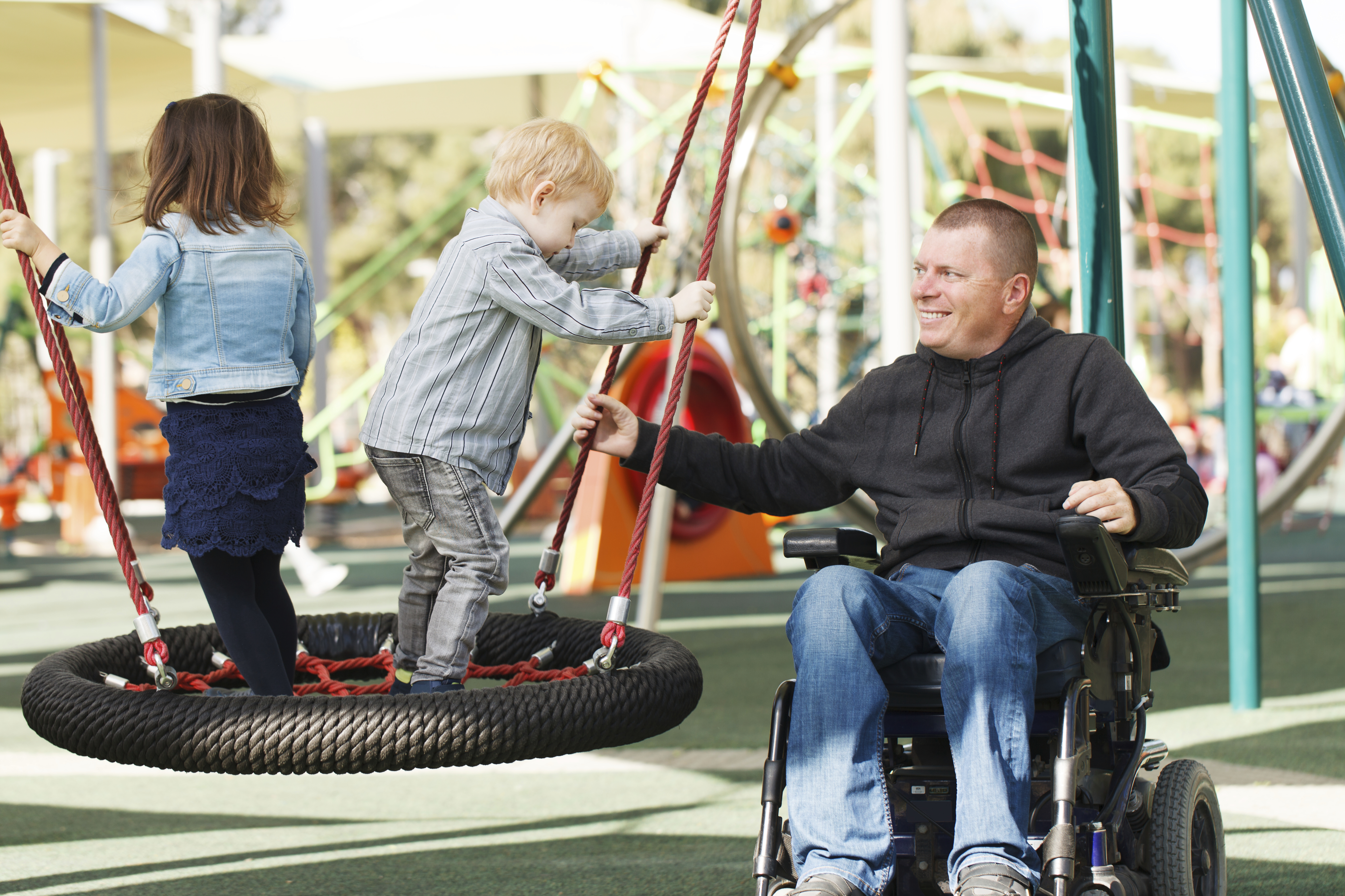 Mann im Rollstuhl auf dem Spielplatz mit seinen Kindern